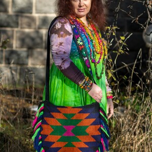 May include: A woman wearing a colorful, patterned dress and a beaded necklace carries a large, fringed bag with a geometric design. The bag is made of woven fabric and has a black strap.