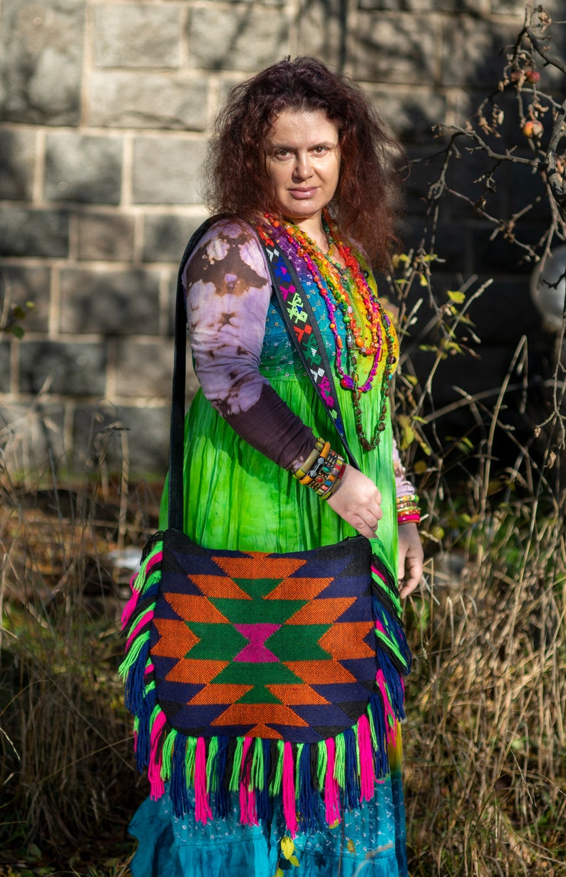 May include: A woman wearing a colorful, patterned dress and a beaded necklace carries a large, fringed bag with a geometric design. The bag is made of woven fabric and has a black strap.