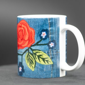 May include: A white ceramic mug with a denim-like design featuring a red rose and green leaves embroidered on the front.