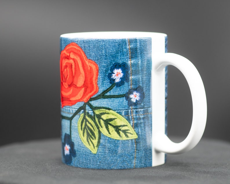 May include: A white ceramic mug with a denim-like design featuring a red rose and green leaves embroidered on the front.