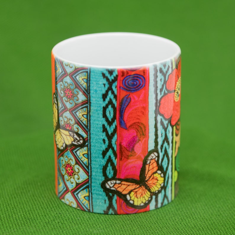 May include: A white ceramic mug with a colorful design featuring butterflies, flowers, and geometric patterns. The design is in shades of blue, red, yellow, green, and black.