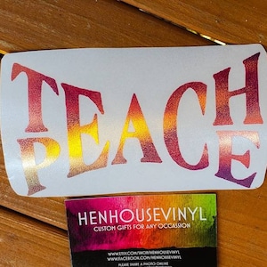 May include: A white vinyl decal with the words "Teach Peace" in a rainbow gradient. The decal is on a wooden surface. The text "HENHOUSEVINYL CUSTOM GIFTS FOR ANY OCCASSION" is on a business card below the decal.