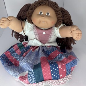 May include: A vintage doll with brown yarn hair styled in pigtails. The doll has a tan face, large eyes, and a small smile. It wears a white top with a pink bib and a patchwork skirt in shades of blue, red, and green. The skirt is trimmed with white lace.