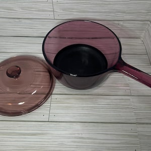 May include: A vintage, purple-tinted glass saucepan with a matching lid. The saucepan has a long handle and a dark interior. The lid is round with a small knob. The set is displayed on a light-colored wooden surface.