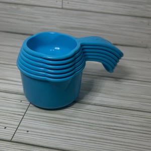 May include: A stack of six light blue measuring cups with handles. The cups are made of plastic and are nested together. The handles are curved and easy to grip. The cups are on a light gray wooden surface.
