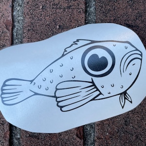 May include: A black and white cartoon fish sticker with a large eye and a smiling expression. The fish has a rounded body with small dots and fins. The sticker is on a white background.
