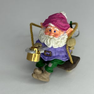 May include: A whimsical gnome figurine on a swing. The gnome wears a purple jacket, green pants, and a pink hat. It has a white beard and is holding a gold-colored object. The swing is suspended by a rope.