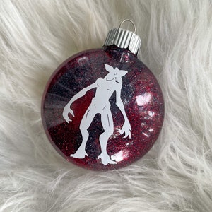 May include: A red and black glitter Christmas ornament with a white silhouette of a monster from the show Stranger Things.