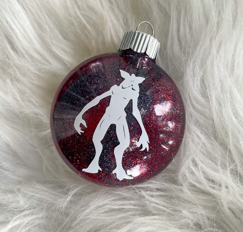 May include: A red and black glitter Christmas ornament with a white silhouette of a monster from the show Stranger Things.