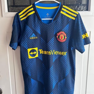 May include: A blue and black Manchester United soccer jersey with yellow accents. The jersey features the Adidas logo, the TeamViewer logo, and the Manchester United crest. The jersey is hanging on a white hanger.