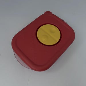 May include: A red plastic container with a yellow circular button in the center. The container has a rectangular shape with rounded corners and a clear, curved edge. The yellow button has two raised circular bumps.