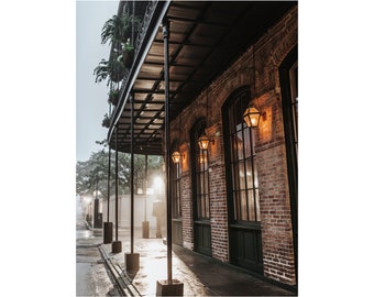 May include: A brick building with a wrought iron balcony and several windows. The windows have black frames and are lit by lanterns. The building is on a street with a sidewalk and a cobblestone road.