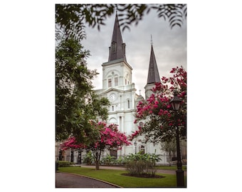 May include: A white church with two tall spires, surrounded by lush green trees and pink flowers. The church has a clock on the front.