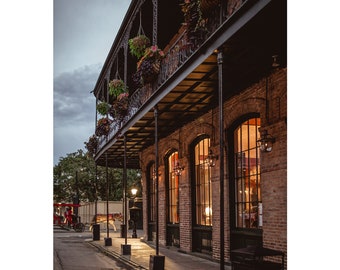 May include: A brick building with a wrought iron balcony and hanging plants. The building has large windows with warm light shining through. A horse-drawn carriage is visible in the street.