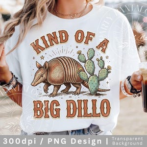 May include: White t-shirt featuring a brown armadillo and green cactus graphic with the text "KIND OF A BIG DILLO" in brown. The design includes stars and sunburst details. The shirt is a casual wear item.