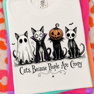 May include: A white t-shirt featuring a graphic of four cats dressed for Halloween: one as a ghost, one as a vampire, one with a pumpkin head, and one as a skeleton. Below the cats, the text reads, "Cats, Because People Are Creepy."
