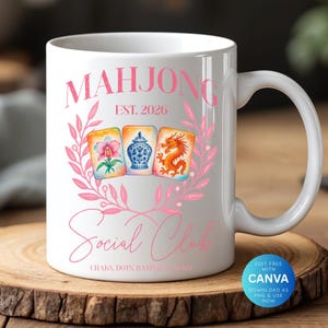 May include: White ceramic mug featuring the words "MAHJONG Social Club" in pink script, with "EST. 2026" above. The design includes a pink floral wreath and watercolor illustrations of Mahjong tiles. The mug sits on a wooden surface.