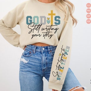 May include: A beige sweatshirt with the text "GOD IS Still writing your story" on the front and "So stop trying to STEAL THE PEN" on the sleeve. The sweatshirt has a cross with hearts on the front.