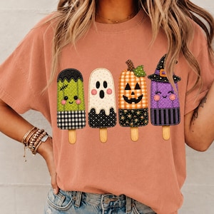 May include: A peach-colored t-shirt featuring a graphic print of four Halloween-themed popsicles. The designs include a green Frankenstein popsicle, a white ghost popsicle, an orange pumpkin popsicle, and a purple witch popsicle with a hat.