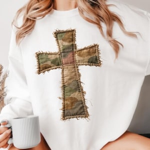 Puede incluir: Sudadera blanca con un diseño de cruz grande de patchwork. La cruz está hecha de tela de camuflaje en tonos verdes, marrones y rosas, con un borde deshilachado. Un estilo casual y cómodo.