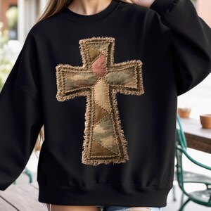 May include: Black sweatshirt featuring a large, textured cross applique. The cross is made of various fabric patches in shades of brown, green, and pink, with a frayed edge. The sweatshirt has a crew neck and long sleeves.