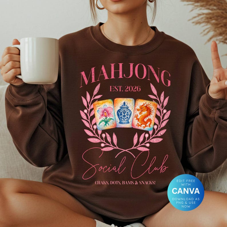 May include: A brown sweatshirt with a pink graphic that reads "MAHJONG Social Club, Est. 2026." The design includes watercolor illustrations of mahjong tiles and a floral wreath. The text "CRAKS, DOTS, BAMS & SNACKS!" is also included.