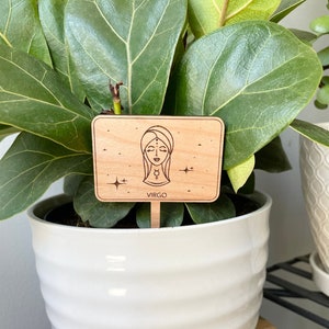 May include: A wooden plant marker with a laser-engraved illustration of a woman's face with a star and the word "VIRGO" below. The marker is in front of a green plant in a white pot.