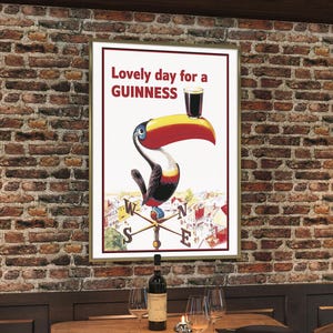 May include: Framed vintage Guinness advertisement featuring a toucan holding a pint of Guinness. The poster reads "Lovely day for a Guinness" in red text. The toucan has a yellow beak, red accents, and is set against a white background. The poster is framed and hangs on a brick wall.