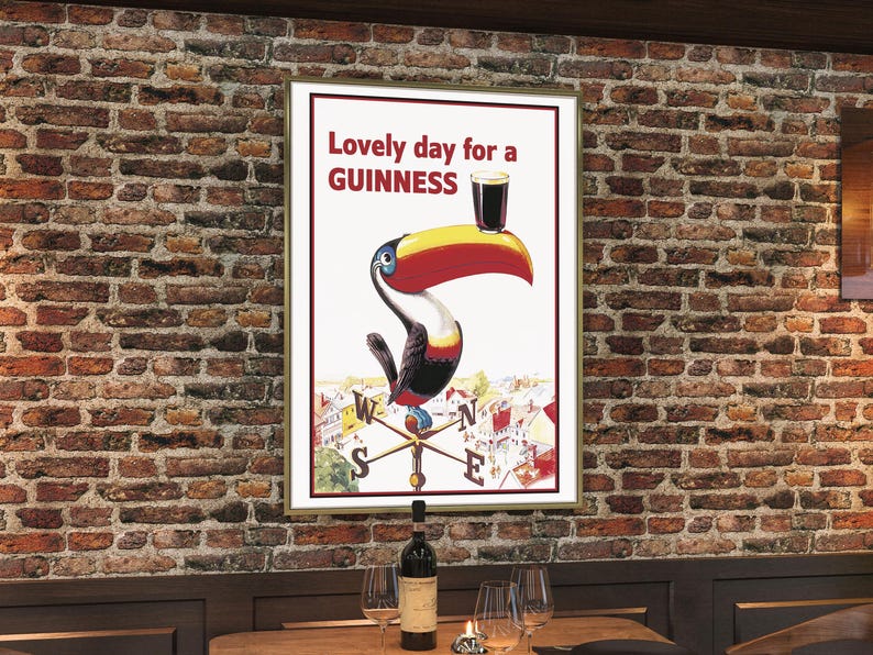 May include: Framed vintage Guinness advertisement featuring a toucan holding a pint of Guinness. The poster reads "Lovely day for a Guinness" in red text. The toucan has a yellow beak, red accents, and is set against a white background. The poster is framed and hangs on a brick wall.