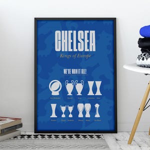 Puede incluir: Un póster azul enmarcado con la palabra "CHELSEA" en letras blancas. Debajo, se lee "Kings of Europe" y "WE'VE WON IT ALL!" con ilustraciones de trofeos. El póster está apoyado contra una pared blanca, junto a una pila de libros y una silla con ropa doblada.
