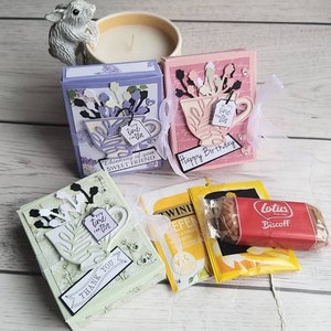 May include: Three decorative gift boxes in lavender, pink, and mint green, each with a teacup design and floral accents. The boxes have ribbons and tags with phrases like "Happy Birthday" and "Thank You." Tea bags and a Lotus Biscoff biscuit are included.