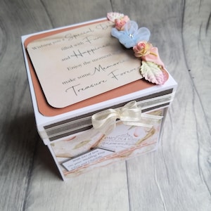 May include: A square gift box decorated with floral accents and a cream bow. The box has text that reads "Wishing you a Special Day filled with Fun and Happiness...Treasure Forever."