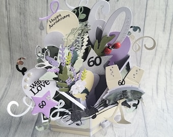 May include: A purple and white 3D pop-up card with the words "Happy Anniversary" and "With Love" and the number "60" on it. The card is decorated with flowers, leaves, and a champagne glass.