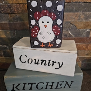Peut inclure: Un bloc de bois peint à la main avec un motif de poulet, surmontant deux autres blocs. Le poulet est blanc avec des accents rouges et des pieds orange, sur un fond brun foncé avec des pois blancs. Le mot "Country" est peint sur le bloc du milieu, et "KITCHEN" en bas.