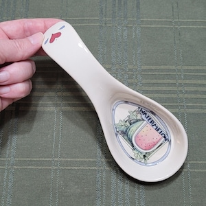 May include: White ceramic serving spoon with a watermelon design. The spoon has a handle shaped like a face with blue eyes and a red heart for a mouth. The watermelon design features a slice of watermelon with green leaves and the word "WATERMELON" written around it.