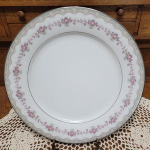 May include: White porcelain plate with a scalloped edge and a floral design. The plate features a delicate border of pink roses and a silver trim. The plate is set on a decorative lace doily.