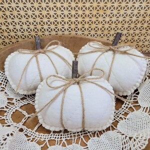 May include: Three white fabric pumpkins with brown stems and twine detailing. The pumpkins feature a quilted pattern and a scalloped edge. They are displayed on a wooden surface with a lace doily.