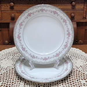May include: Two white porcelain plates with a floral design. The plates have a silver rim and a decorative border with pink flowers and a grey background. One plate is on a clear stand.