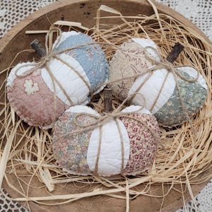 May include: Three fabric pumpkins crafted from quilted panels in assorted floral and solid white patterns. Each pumpkin features a small, dark brown twig stem and is wrapped with thin, natural-colored twine tied in a bow at the top.