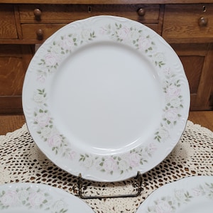 May include: A set of three white ceramic dinner plates with a delicate floral design featuring pink roses and green leaves. The plates have a slightly scalloped edge and a gold rim.
