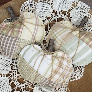 May include: Three patchwork fabric heart-shaped ornaments tied with twine, in shades of brown, green, and cream, displayed on a wooden bowl with a white crocheted doily.