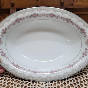 May include: Oval serving dish with a white interior and a decorative border. The border features a scalloped edge with pink floral accents and a silver trim. The dish has handles with a brown and silver design.