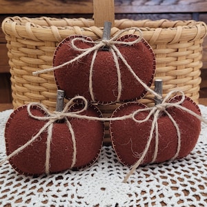 May include: Three dark orange felt pumpkins with visible stitching along the edges. Each pumpkin features a small, natural wooden twig stem and is wrapped with thin, light brown jute twine tied into a simple bow at the top of the stem.