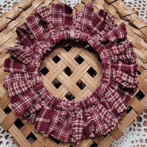 May include: A circular, ruffled wreath with a burgundy and cream plaid pattern. The wreath is made of fabric and has a frayed edge. It sits on a woven, wooden surface.