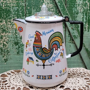 May include: A white enamel coffee percolator featuring a colourful rooster illustration, a black handle, and a clear glass knob on the lid. The design includes the text Good Morning, Wake up, and Coffee time, along with small house and flower motifs.