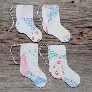 Könnte beinhalten: Vier kleine, gesteppte Weihnachtsstrümpfe mit weißen Schnurschlaufen zum Aufhängen. Jeder Strumpf besteht aus Patchwork-Stoff in verschiedenen Pastellfarben, darunter Rosa, Blau und Grün, mit Blumen- und Vichy-Mustern. Die Strümpfe sind auf einer Holzoberfläche angeordnet.
