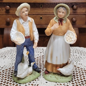 May include: Two ceramic figurines depicting a farmer and his wife. The farmer wears a straw hat, vest, and holds a basket. His wife wears a bonnet and apron, also holding a basket. A goose and chicken are at their feet.
