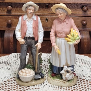 May include: A pair of ceramic figurines depicting an elderly couple. The man wears a white shirt, red vest, and gray pants, holding a hoe and a basket of potatoes. The woman wears a pink shirt, white apron, and blue skirt, holding corn and a baby rabbit.