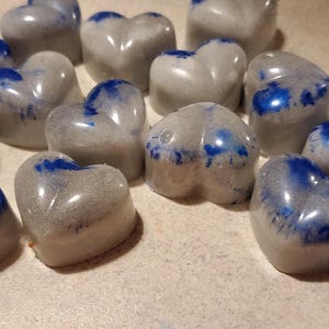 May include: A collection of small, heart-shaped wax melts featuring a smooth, light gray base with vibrant, swirled blue accents. Each individual heart has a glossy finish and a rounded, three-dimensional shape, scattered across the frame.