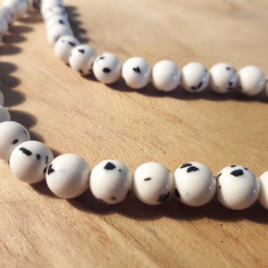 May include: A strand of white beads with black speckles. The beads are round and smooth.